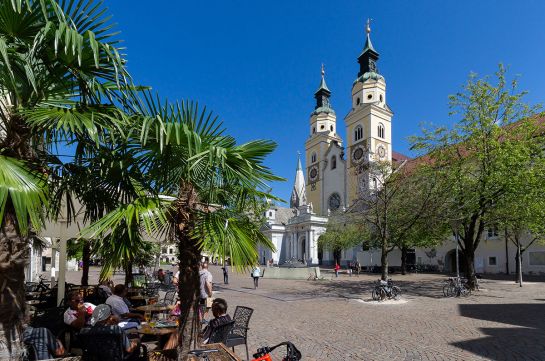 Fotografie - Partnerstadt Brixen - Menschen sitzen im Cafe, im Hintergrund großer Platz mit Kirche