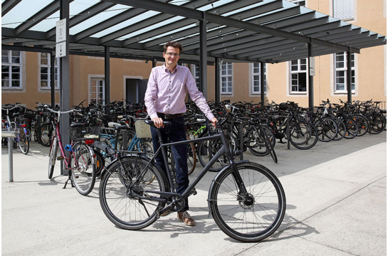 Fotografie: Thomas Großmüller steht mit seinem Fahrrad im Innenhof des Neuen Rathauses. Im Hintergrund sind Fahrradständer mit vielen Fahrrädern zu sehen. 
