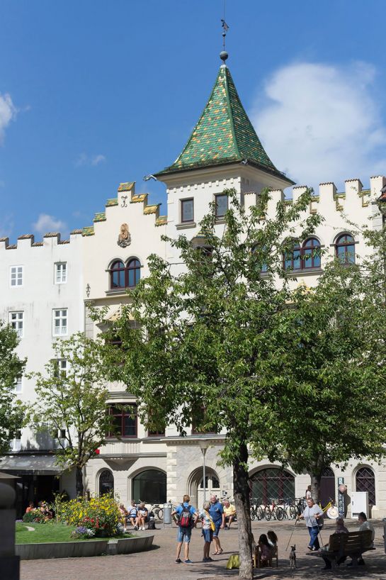 Fotografie - Partnerstadt Brixen - Platz mit Bäumen und Sitzgelegenheiten, im Hintergrund Häuserzeile mit Turm
