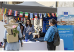 Informationsstand der Europäischen Kommission dekoriert mit kleinen Fahnen der EU-mitgliedsstaaten. Junge Frau mit Sonnenbrille spricht mit einer Besucherin. Eine weitere Besucherin beobachtet die Szene.