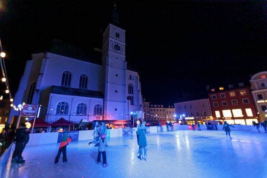 Menschen auf Schlittschuhen auf einer beleuchteten Eisfläche. Im Hintergrund eine Kirche
