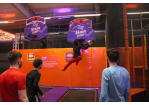 Jugendlicher in der Trampolinhalle springt mit einem Basketball in der Hand vor dem Basketballkorb auf dem Trampolin. 3 Freunde stehen im Vordergrund des Bildes und beobachten ihn.