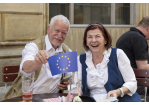 älteres Ehepaar sitzt an einem runden Biergartentisch. Der Mann zeigt eine kleine Europa-Flagge in die Kamera.