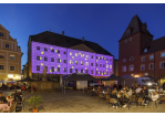 Am Haidplatz in der Regensburger Innenstadt werden auf die Fassade des Thon-Dittmer-Palais  EU-Motive mit blauem Hintergrund und gelben Sternen projiziert. Auf dem Platz sind rund um den Brunnen Pavillons zu sehen. Viele Personen nutzen das Angebot der Außengastronomie