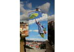 Fotografie – Bild an einer Hausfassade, Eine ältere Dame mit fliegt mit aufgespannten grünen Regenschirm in einen, mit wenigen Wolken, blauen Himmel
