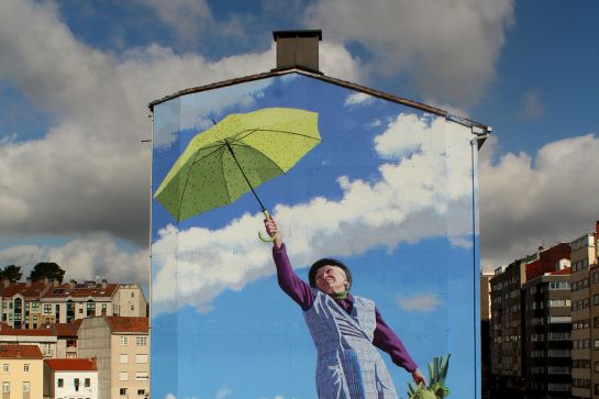 Ein bemaltes Haus mit Himmel und Stadtlandschaft im Hintergrund. Die Mural zeigt eine alte Dame, die mit ihrem Regenschirm gen Himmel fliegt.