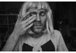 Eine Fotografie in Schwarz-Weiß mit Portrait des Künstlers Angela Aux mit Wu-Tang-Shirt und blonder Perücke, den Blick gesenkt, das rechte Auge durch Finger seiner Hand verdeckt.