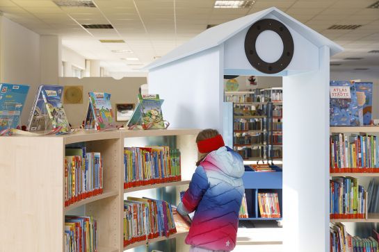 halbkreisförmiges Bücherregal mit Kinderbüchern. Ein Mädchen sucht ein Buch daraus aus