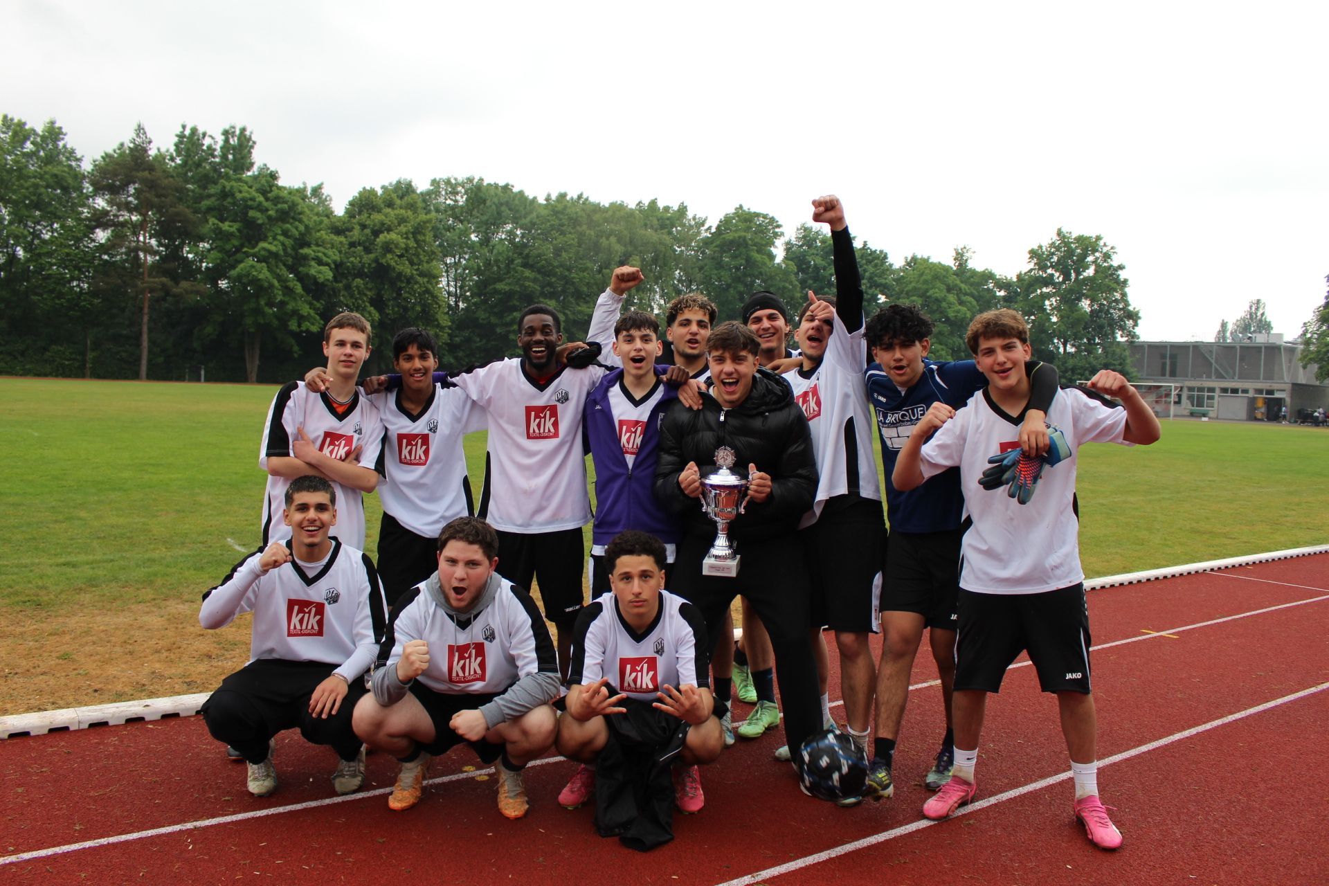Auf dem Bild sind 14 Jugendliche in Sportkleidung sowie ihr jugendlicher Trainer in einer schwarzen Jacke zu sehen. Die Spieler in der Mitte halten den Pokal in die Luft. Das Bild entstand im Mai 2025 beim JUZ übergreifenden Fußballturnier