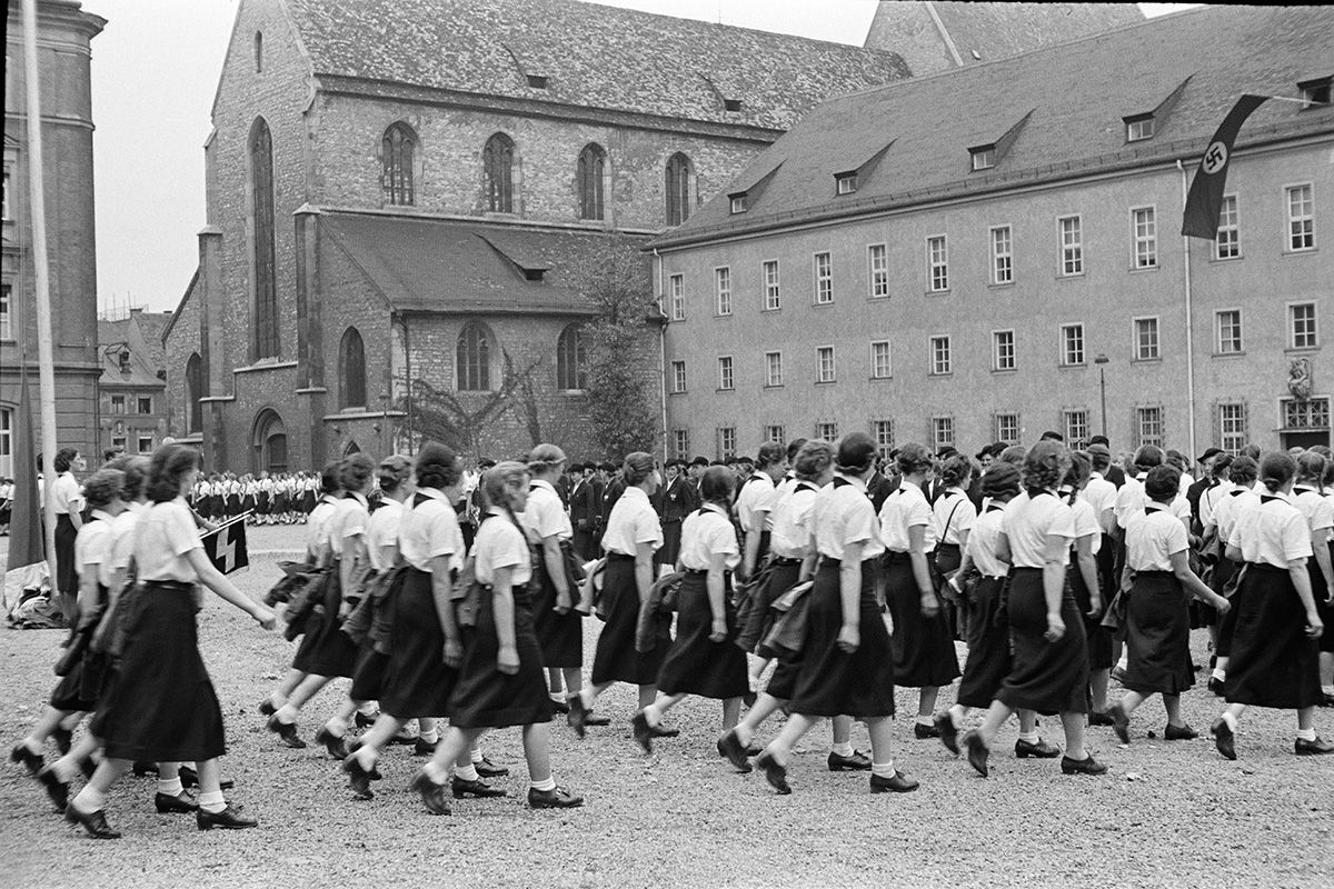 Zu sehen sind Mädchen vom Bund deutscher Mädel, die bei einer Parade im Gleichschritt marschieren. Die Mädchen tragen alle lange dunkle Röcke, schwarze Schuhe, weiße Socken und weiße Hemden. Im Hintergrund ist eine NS-Fahne zu sehen.