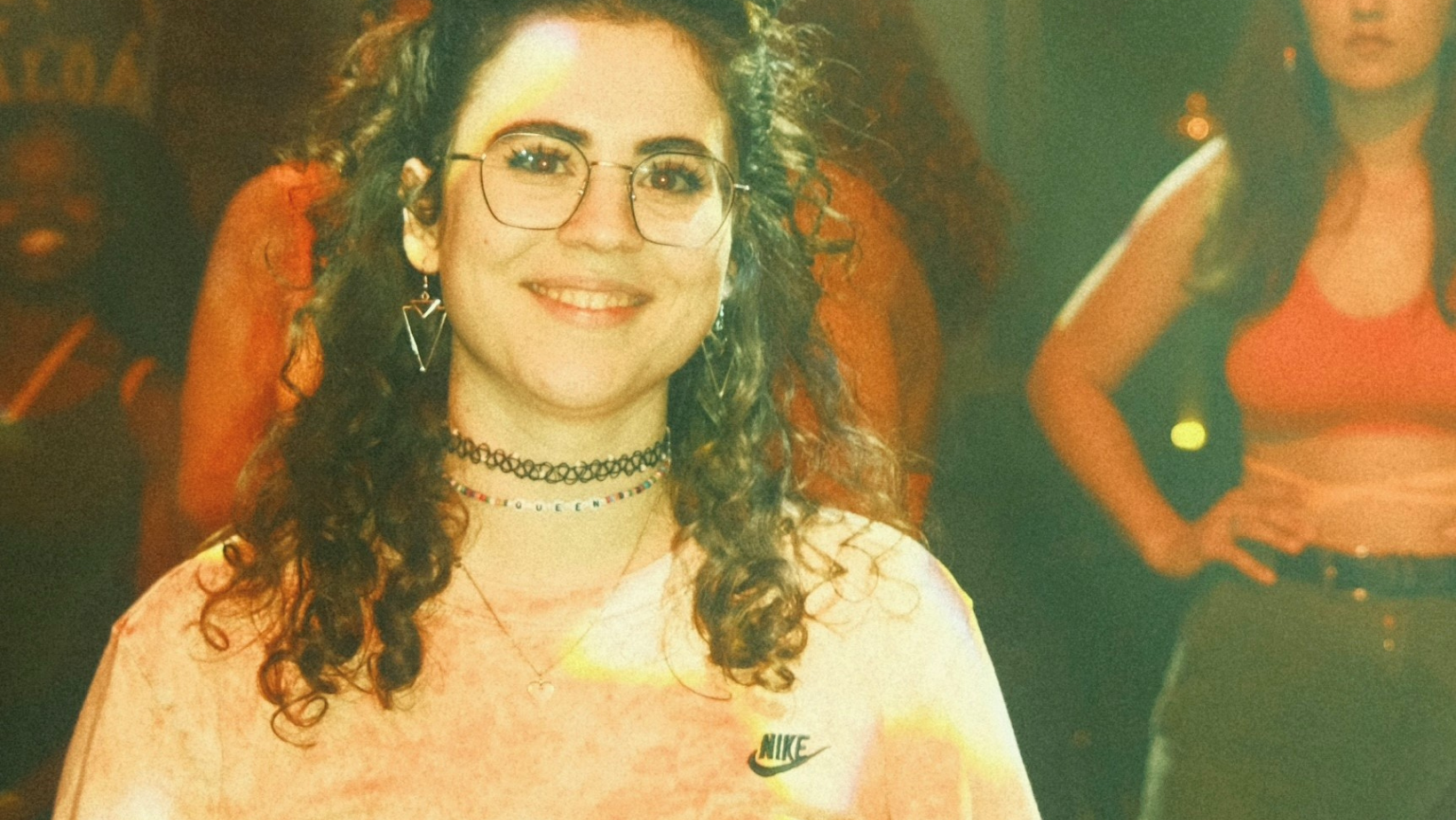 Auf dem Foto ist Regina Schweiger, die den Dancehallkurs im W1 leitet, zu sehen. Sie lächelt dabei in eine Kamera, im Hintergrund sind Menschen zu sehen und das Bild hat einen Analogen Look.