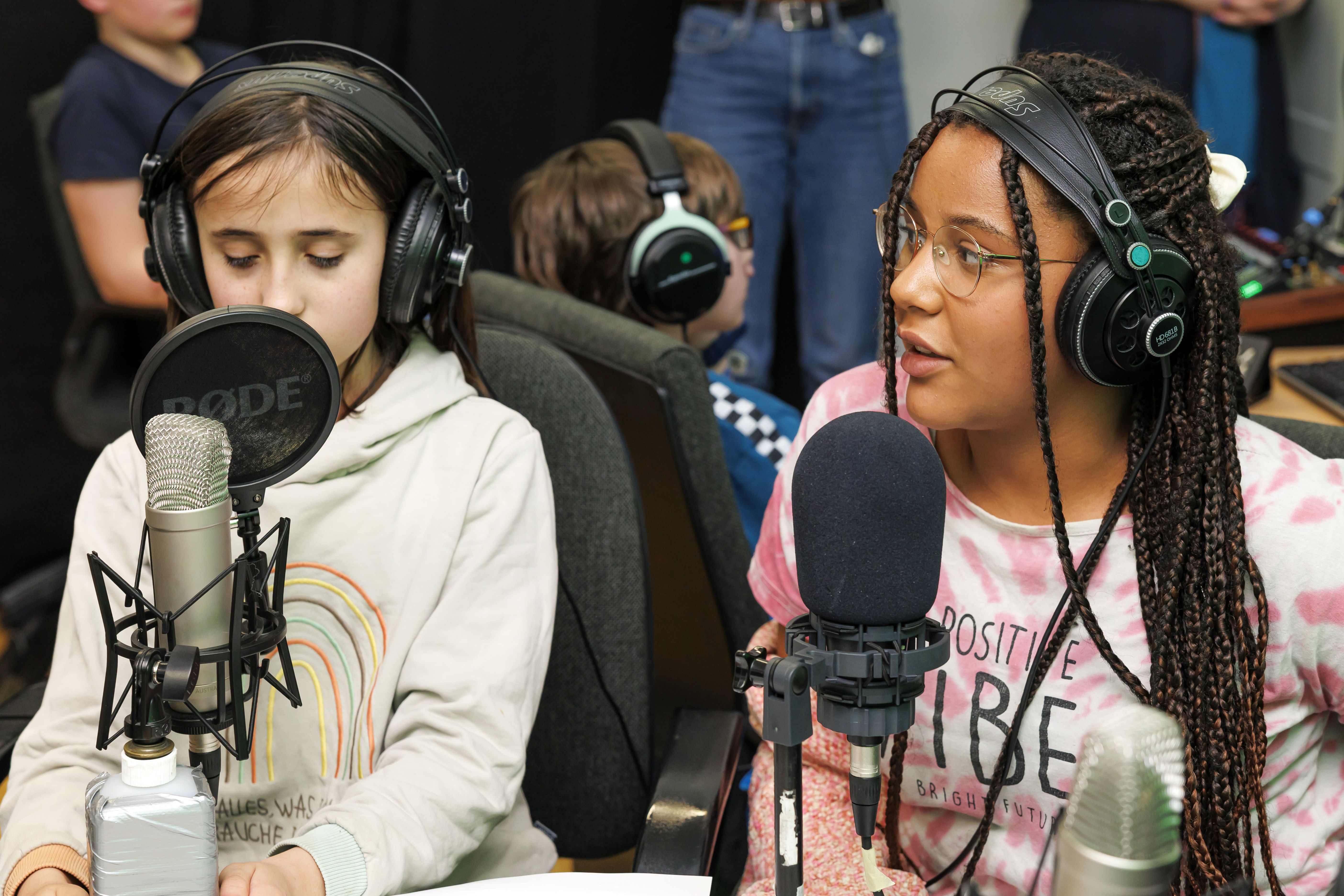 Zwei Mädchen sitzen vor zwei Podcast-Mirkofonen. Im Hintergrund sitzt ein Junge am Mischpult. Alle tragen Kopfhörer.