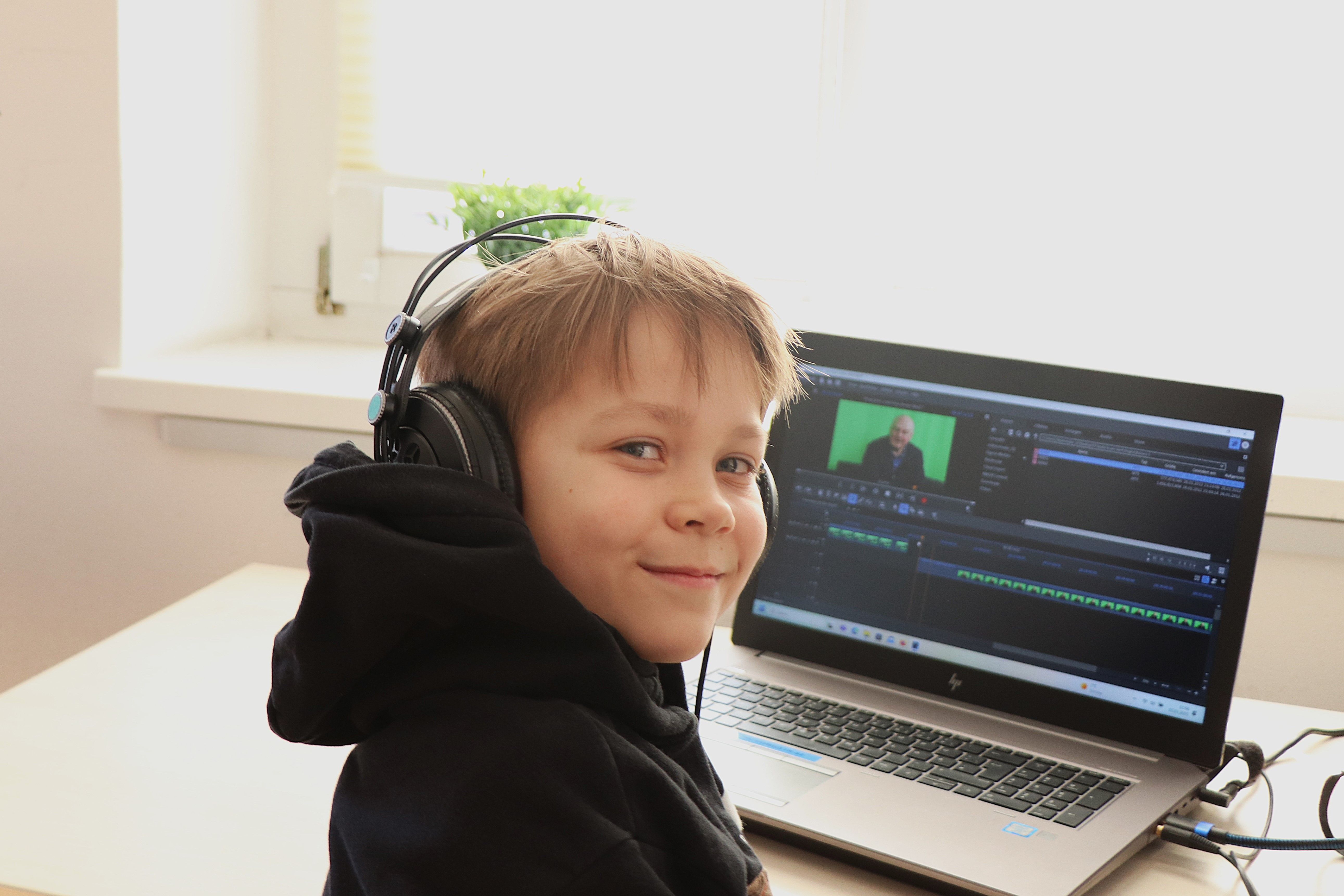 Ein Junge blickt in die Kamera. Er trägt Kopfhörer. Hinter ihm ist ein Laptop, auf dem ein Schnittprogramm läuft.