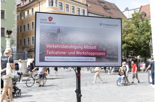 Fotografie: Das Foto zeigt einen auf dem Domplatz aufgebauten Bildschirm mit einer Präsentation mit dem Titel „Verkehrsberuhigung Altstadt - Teilnahme- und Workshopprozess“.