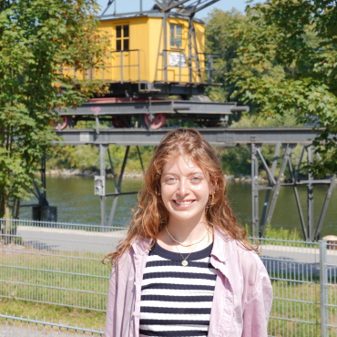 In der Mitte steht ein junges Mädchen mit roten Haaren und einer rosanen Bluse. Sie steht vor der Donau; im Hintergrund hängt ein gelber Krahn.
