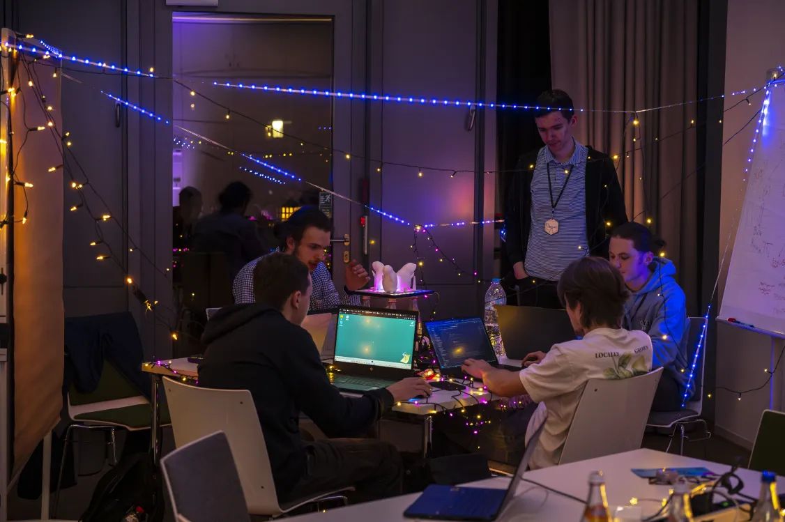 In einem dunkel beleuchteten Raum sitzt ein Team in der Mitte an einem Tisch und arbeitet an den Laptops. Über ihnen hängen Lichterketten.