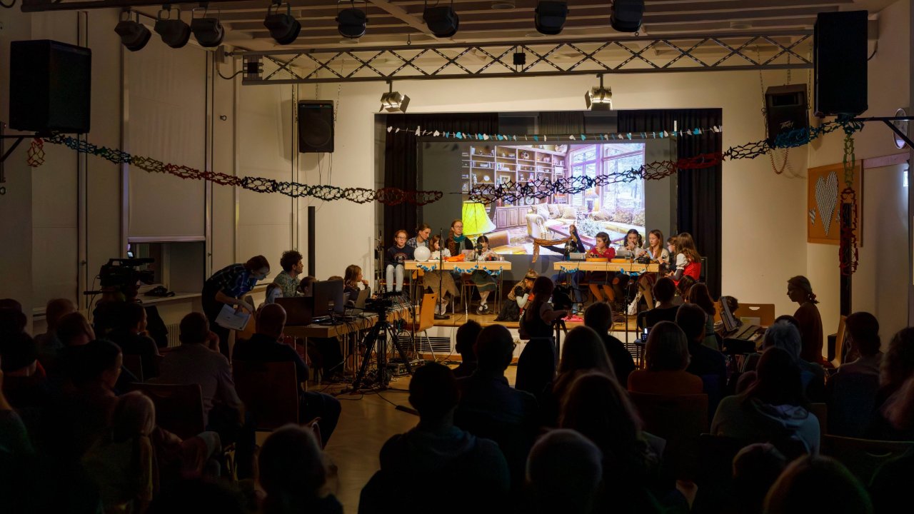 Saal mit vielen Menschen, die auf eine Bühne schauen, auf der Kinder sitzen