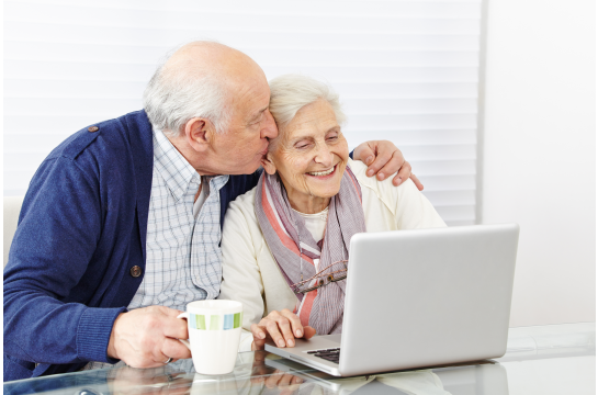 Fotografie: Ein Seniorenpaar sitzt vor einem Laptop. Der ältere Herr hat einen Arm um die Dame gelegt und gibt ihr ein Küsschen.