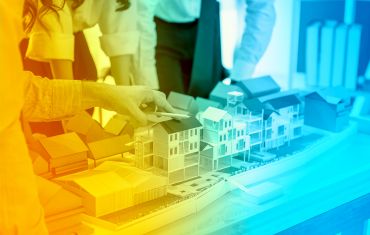 Das Foto hat einen über das Motiv gelegten Farbverlauf. Dieser beginnt links in einem hellen Orangeton, der zur Mitte des Bildes schwächer wird und mittels eines Übergangs in einem leichten Grünton in ein transparents Blau mündet. Das Motiv zeigt die Oberkörper dreier Personen, die über ein Wohngebiet-Modell gebeugt stehen. Die ganz links im Bild erkennbare Person, berührt eines der Häuser des Modells und hält dabei einen Kugelschreiber in der Hand.