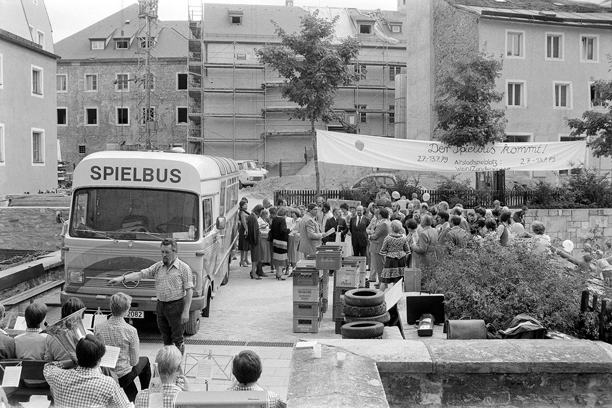 Zu sehen ist der neue Spielbus im Jahr 1979. 
Auf der linken Seite des Fotos ist ein Mercedesbus zu sehen, auf dessen front „Spielbus“ steht. Im Vordergrund ist eine Blaskapelle zu sehen. Im Hintergrund versammeln sich zahlreiche Menschen unter einem Banner mit der Aufschrift „Der Spielbus kommt!“ Ein Mann mit Papieren in der Hand hält eine Rede. Zwischen der Menschenansammlung und der Kapelle stehen die Spielutensilien des Spielbusses.                 