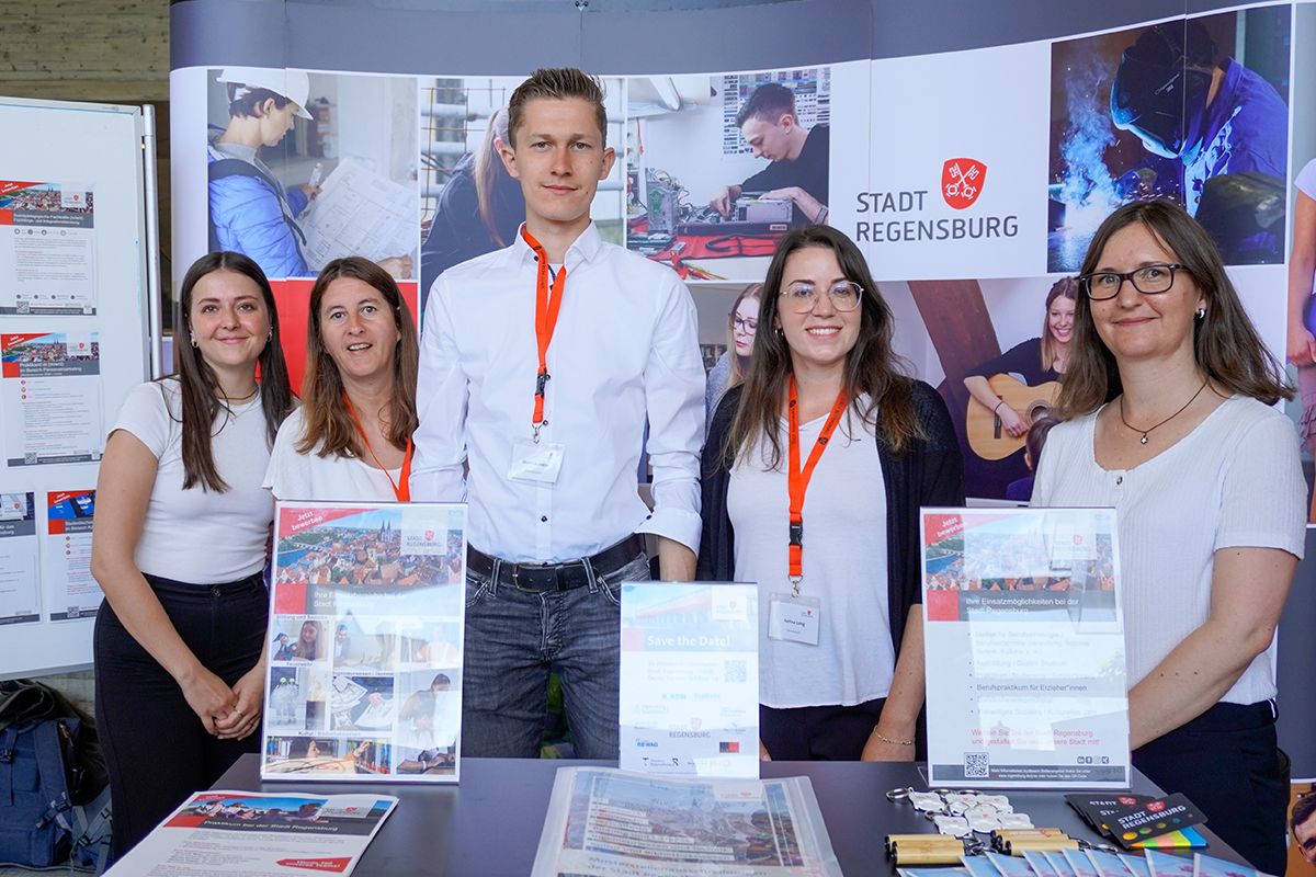: Mehrere Personen posieren an einem Messestand, an dem Flyer und Displays zum Thema Ausbildung und Karriere präsentiert werden.