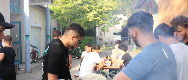 Eindrücke vom Sommerfest des Jugendzentrum Arena. Viele Jugendliche sind zu sehen. Im Vordergrund überreicht ein junger Mann einem anderen einen Burger und im Hintergrund sitzen mehrere junge Männer an einem Tisch und stoßen mit Spezi-Flaschen an