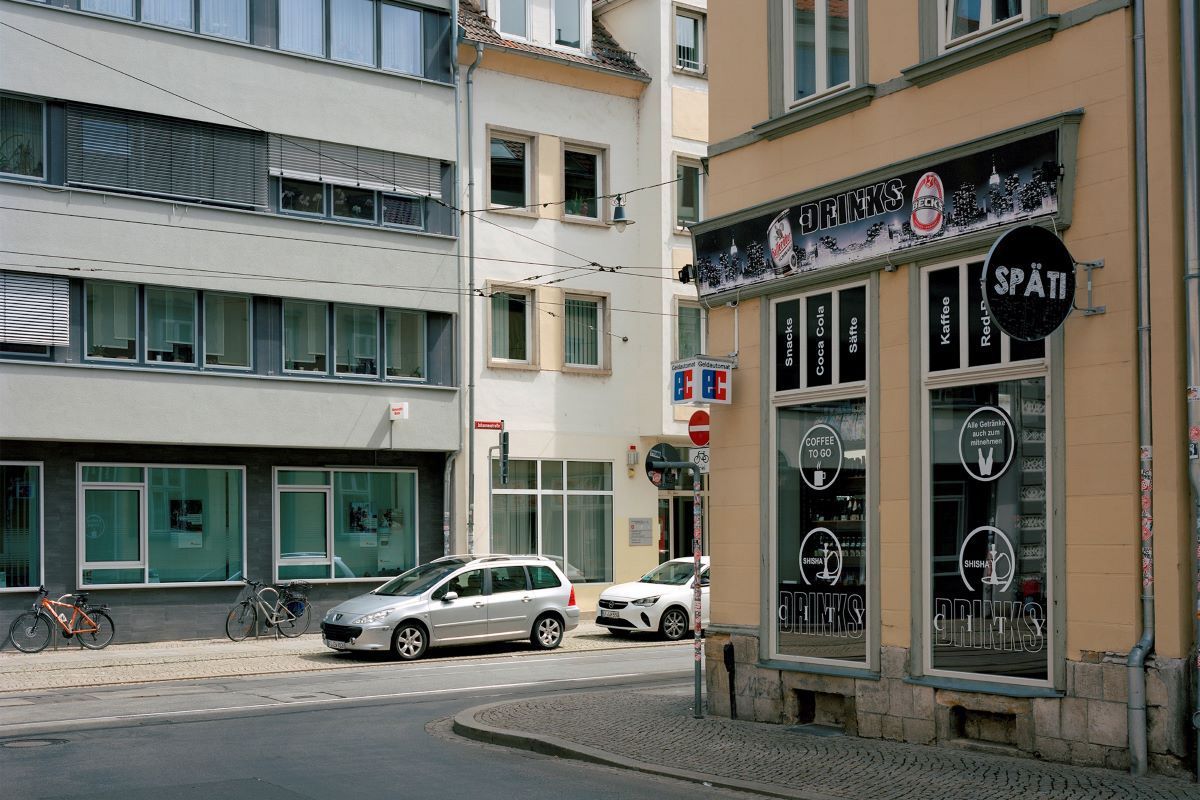 Fotografie - Straßenecke mit Späti auf der rechten Seite, links 2 Gebäude mit parkenden Autos