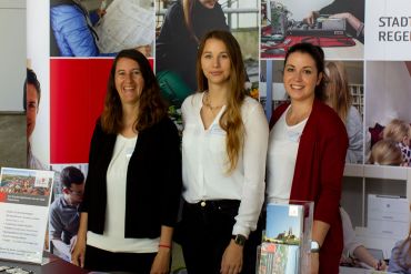 Fotografie – Messestand der Stadt Regensburg mit Vertreterinnen des Personalamts – im Hintergrund eine Messewand mit Fotos von Beschäftigten der Stadt Regensburg