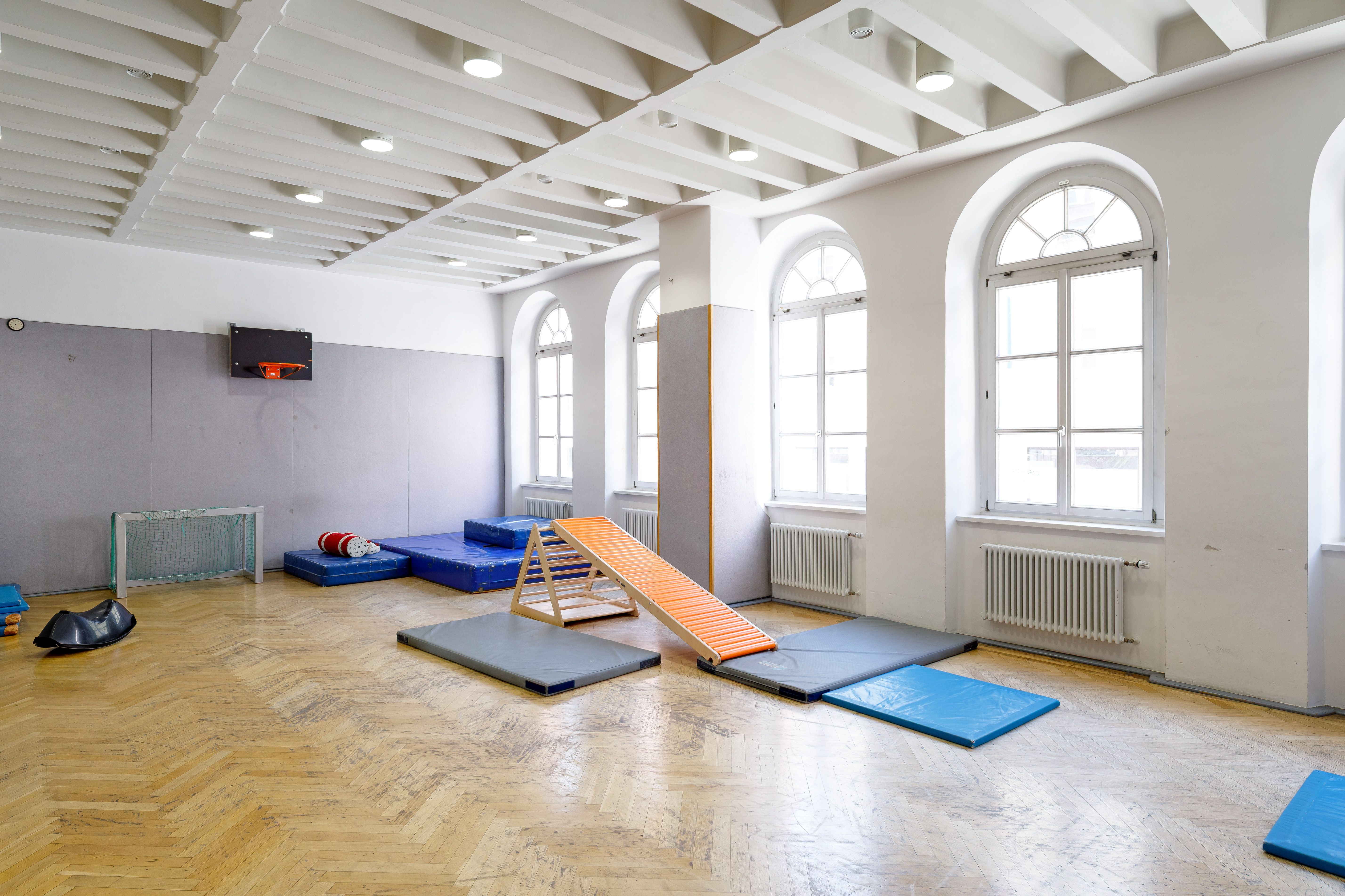 Turnhalle mit Parkettboden und gepolsterter Wand. An der Wand hängt ein Basketballkorb und es liegen verschiedene Matten im Raum verteilt. Auch eine Rollenrutsche und Fußballtore sind zu sehen