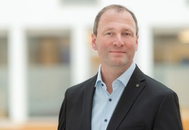 Man erkennt einen Mann im Anzug, der in die Kamera lächelt.