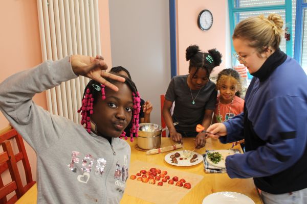 Das Bild wurde in der Küche des Jugendzentrums aufgenommen. Auf dem Tisch in der Mitte sieht man halbierte Erdbeeren auf einem Backpapier. Eine Mitarbeiterin des Jugendzentrums verziert die Erdbeeren gemeinsam mit drei Mädchen mit Schokolade. Im Vordergrund zeigt ein junges Mädchen ein Peacezeichen in die Kamera.