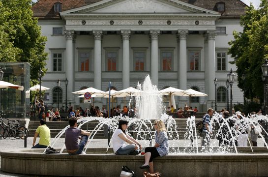 Fotografie - Personen sitzen am Brunnen am Bismarckplatz - im Hintergrund Gebäude Haus der Musik