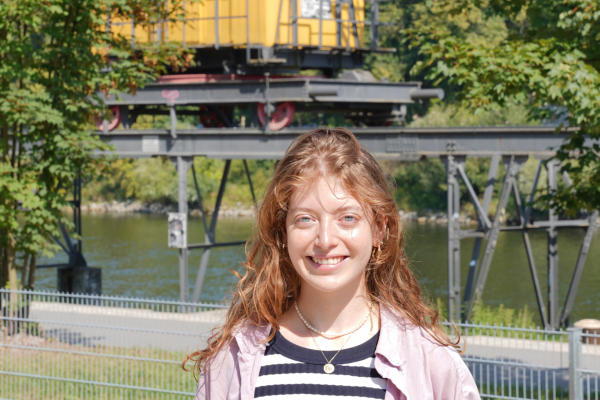 In der Mitte steht ein junges Mädchen mit roten Haaren und einer rosanen Bluse. Sie steht vor der Donau; im Hintergrund hängt ein gelber Krahn.