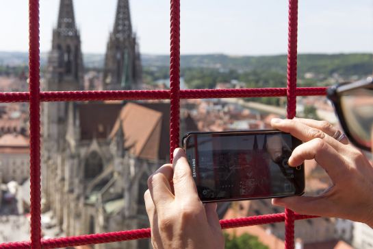 Fotografie: Zwei Hände machen durch ein rotes Gitter ein Handyfoto vom Dom. (C) Bilddokumentation Stadt Regensburg