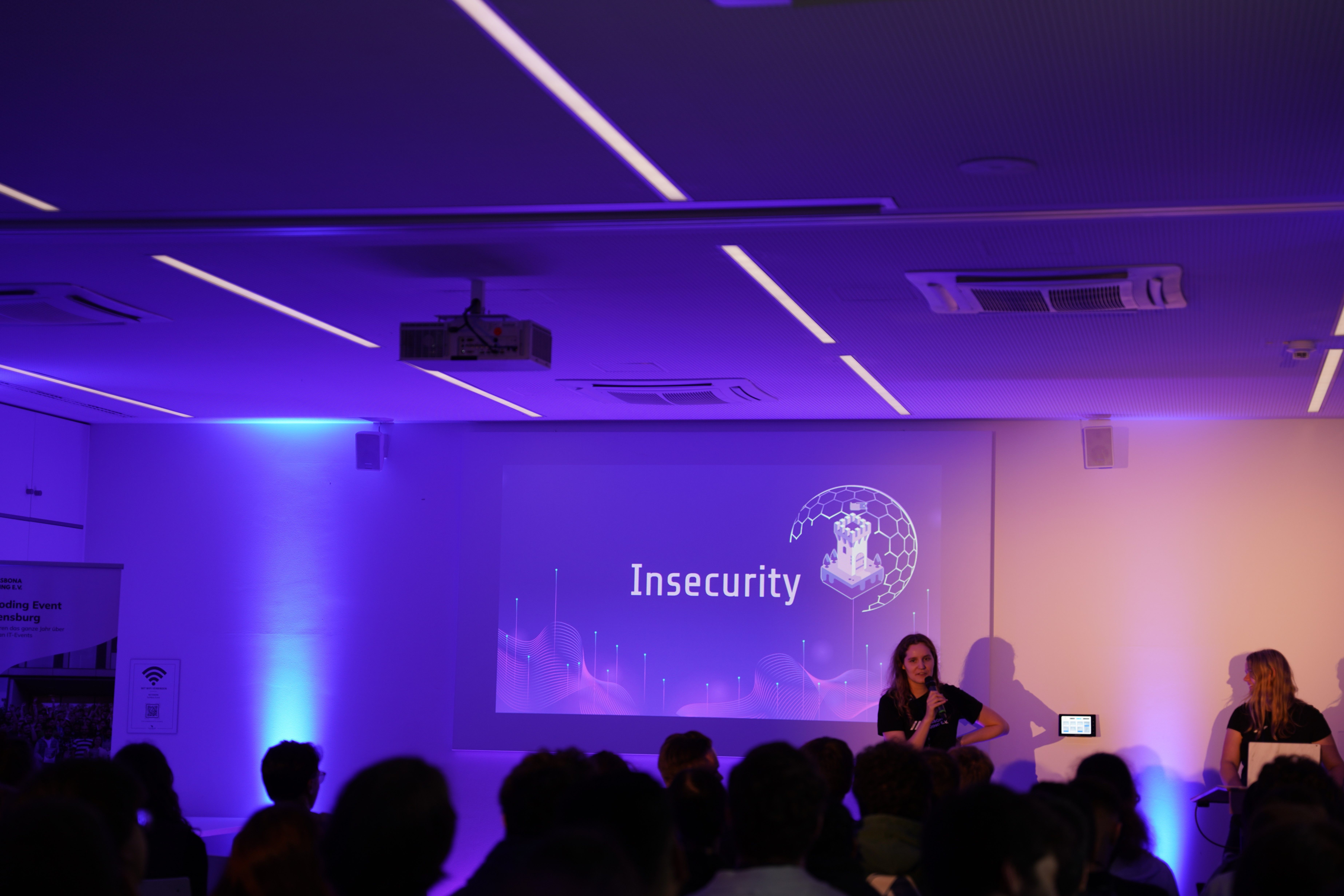 In einem dunkel beleuchteten Raum wird vor einem großen Publikum das Motto "Insecurity" auf einer Leinwand vorgestellt.