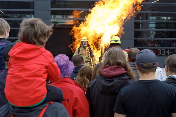 Fotografie: Zwei Feuerwehrler stehen vor einem Feuer, im Vordergrund stehen Zuschauer der Vorführung.