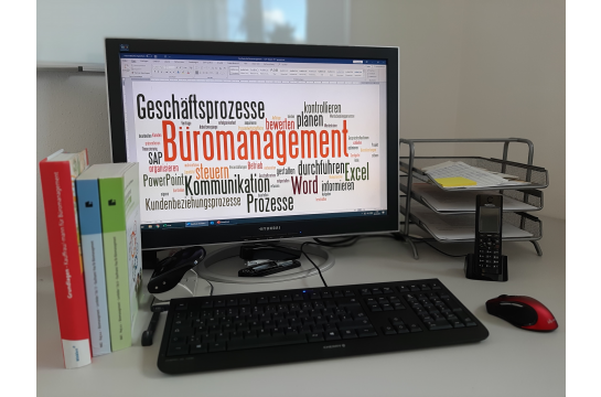 Schreibtischoberfläche mit typischer Büroausstattung inklusive PC-Monitor, der eine Wortwolke mit den Ausbildungsinhalten zeigt