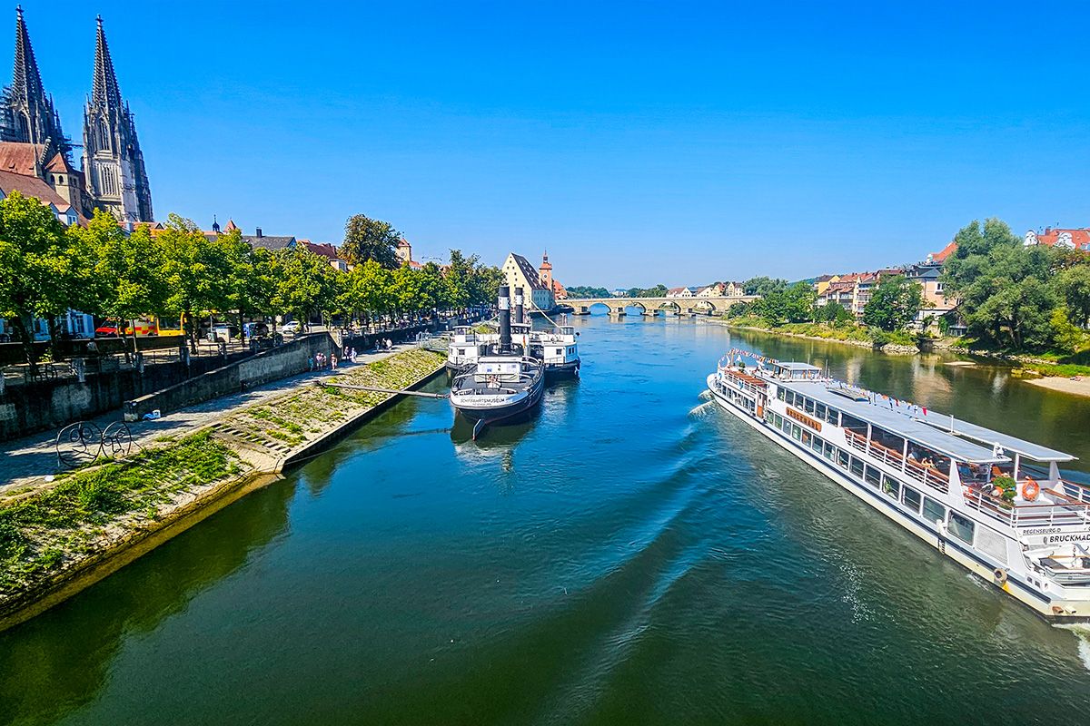 Fotografie - Blick über die Donau auf Regensburg mit Ausflugsschiffen, Uferpromenade und dem Regensburger Dom im Hintergrund bei blauem Himmel.