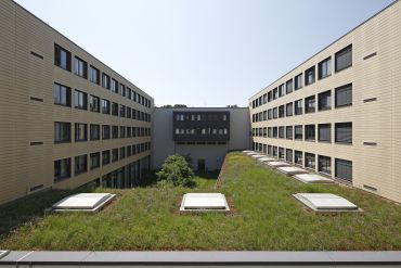 Das Bild zeigt ein begrüntes Flachdach mit mehreren Dachluken. An den Seiten und im Hintergrund sind weitere flache Gebäude zu sehen, die um einen begrünten Innenhof gruppiert sind.