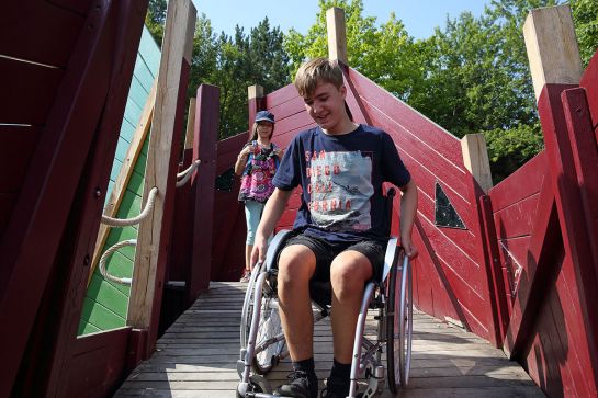 Fotografie – ein Junge fährt im Rollstuhl auf einem Spielgerät des Inklusionsspielplatzes bergab, im Hintergrund ein Mädchen mit blauer Kappe (C) Bilddokumentation Stadt Regensburg