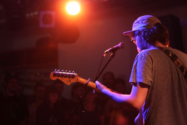 Auftritt der Band "Burkini Beach": Sänger mit Gitarre vor dem Mikrofon, Blick von hinten gegen das Licht eines Scheinwerfers.
