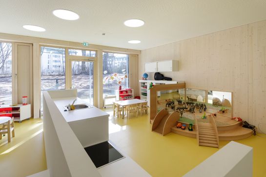 Fotografie - Blick in einen Raum des Kinderhauses,  rechts im Bild eine Spielecke, in der Mitte eine kleine Küchenzeile, im Hintergrund eine große Glasfront