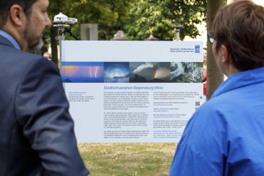 Fotografie: Zwei Personen stehen vor einem Schild zur Stadtklimastation Regensburg-Mitte.