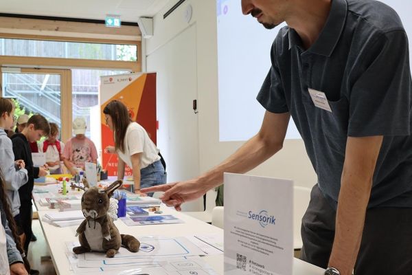 Vom rechten unteren Bildrand zieht sich ein langer Tisch bis zum oberen linken Bildrand. Dahinter steht ein Mann und eine Frau, die sich über den Tisch mit interessierten Kindern über das Thema Sensorik unterhalten.