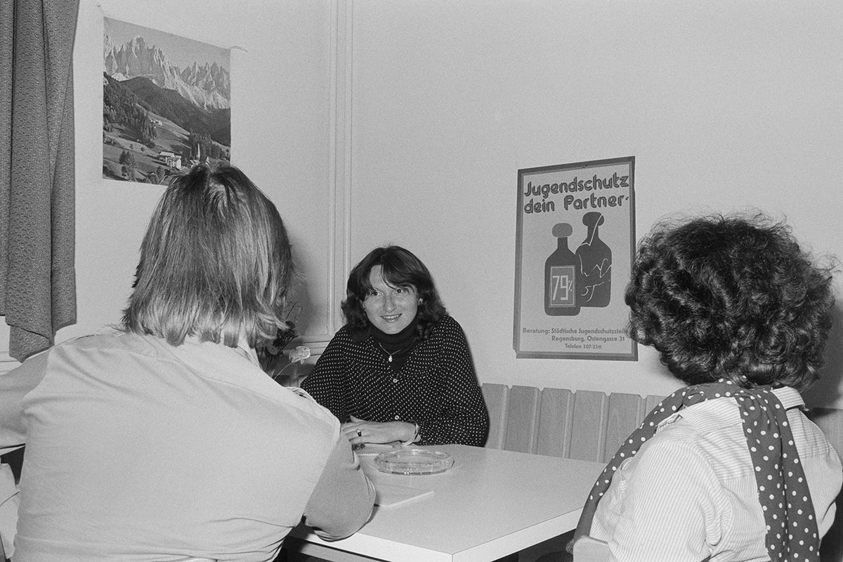 Zu sehen ist eine Beratungssituation in der Erziehungsberatungsstelle um 1977, bei der drei Personen am Tisch sitzen. Die beratende Person, dessen Gesicht zu sehen ist, sitzt den zu beratenden Personen gegenüber. An der Wand hängt ein Plakat, bei dem der Jugendschutz beworben wird.