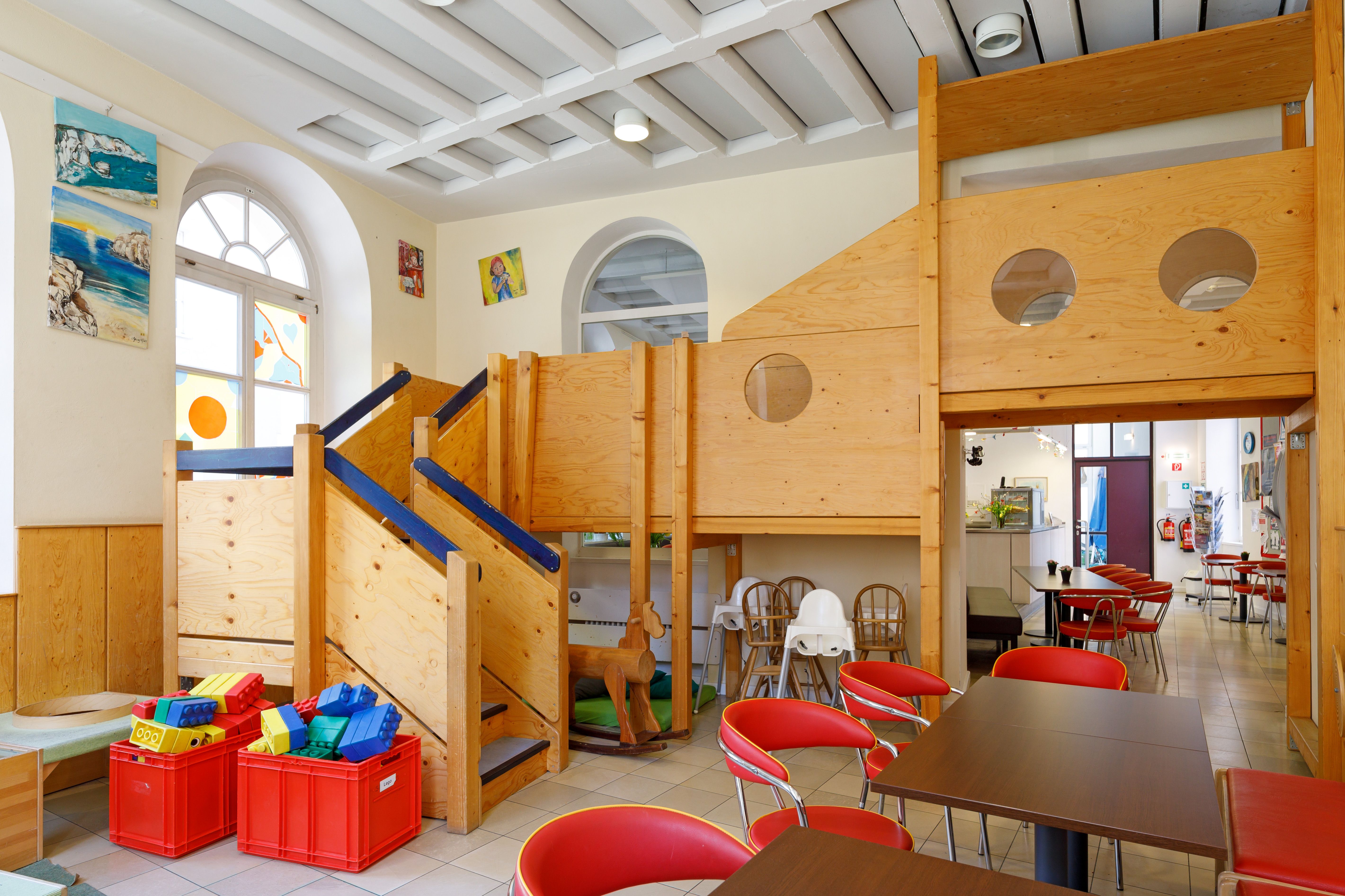 Kletterturm aus Holz mit Bullaugen für Kinder zum durchsehen. große Lego Steine und Hochstühle sowie eine Sitzecke mit Tischen für Gäste sind zu sehen. 