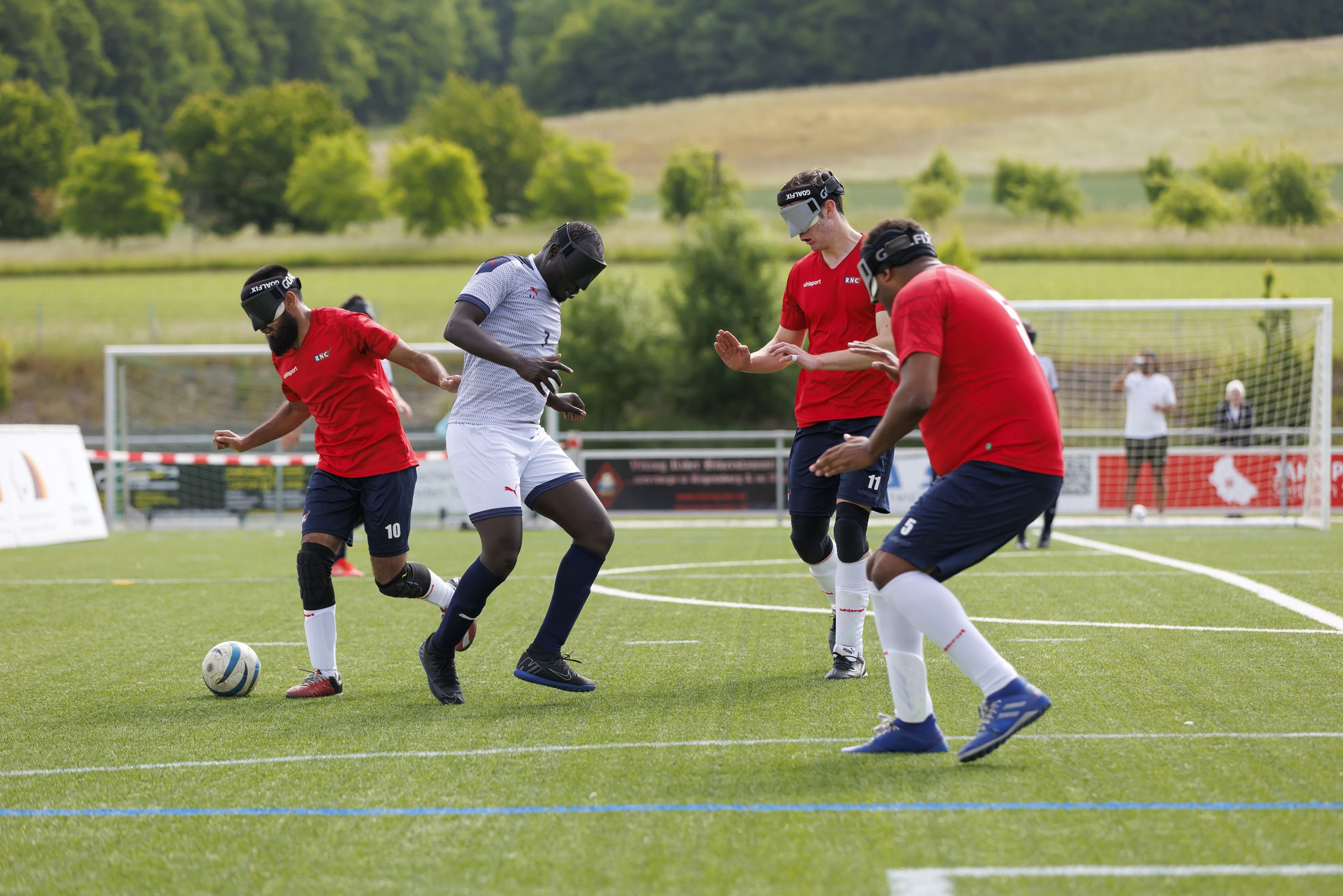 Fotografie: Drei Spieler der englischen Blindenfußball-Nationalmannschaft in roten Trikots und ein Spieler der französischen Blindenfußball-Nationalmannschaft im weißen Trikot bei einem Spiel im Rahmen des Drei-Länder-Cups 