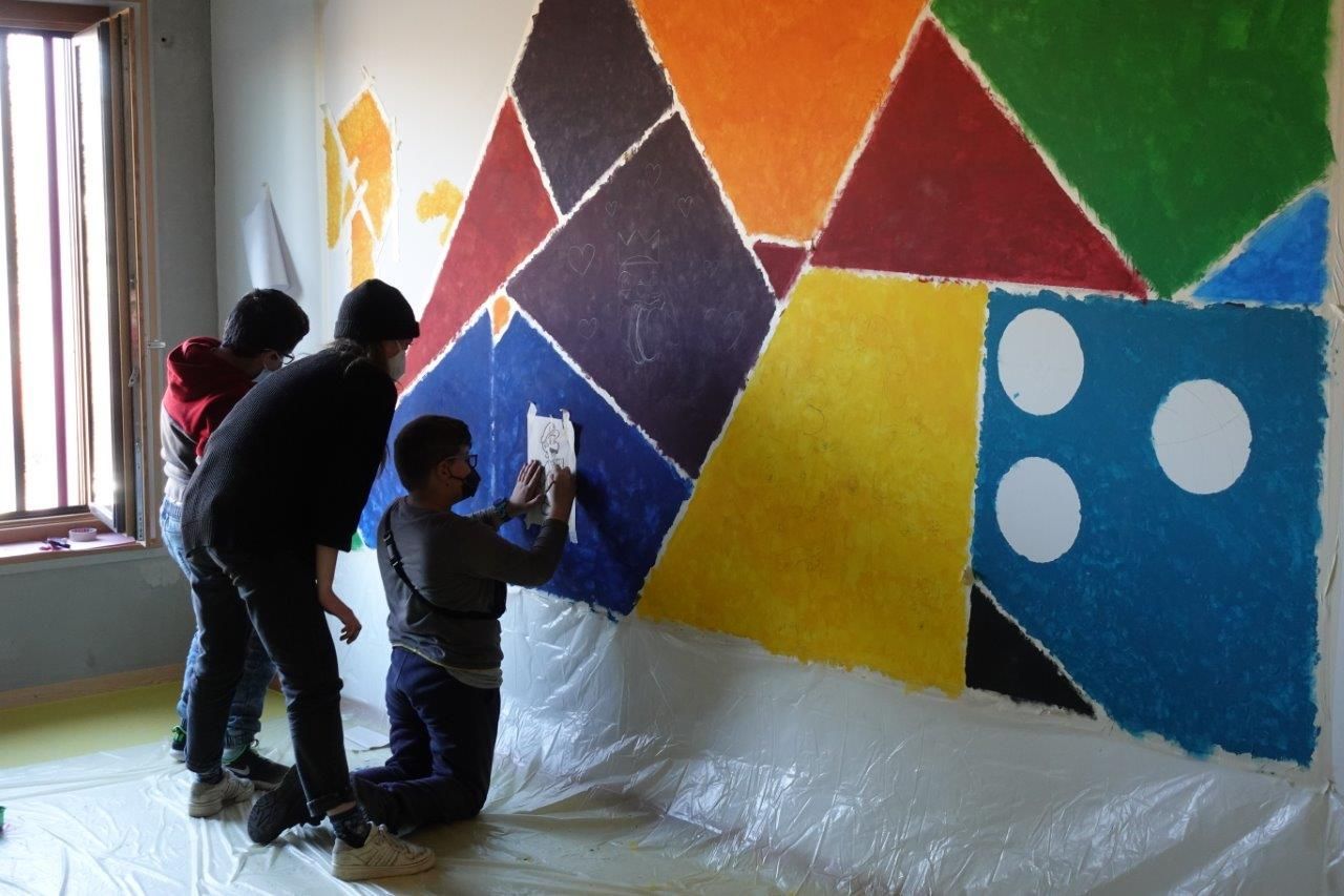 Auf dem Foto sind zwei Kinder und eine erwachsene Person zu sehen welche an der Wand ein Bild malen.