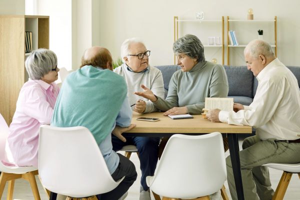 Fotografie: Fünf Seniorinnen und Senioren sitzen gemeinsam mit einem Buch am Tisch und reden.