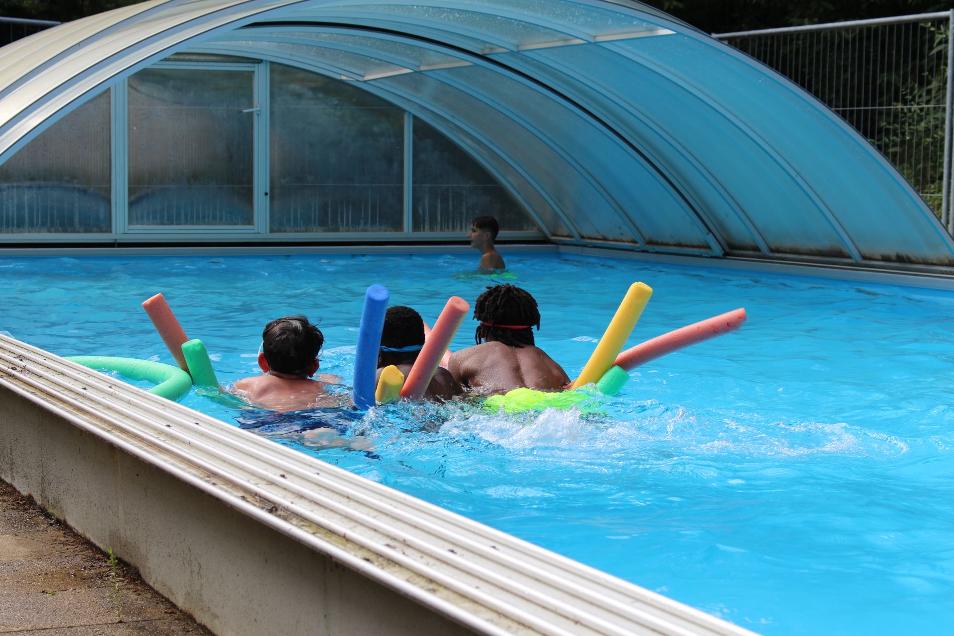 Auf dem Bild ist ein Pool zu sehen. Im Zentrum sind drei Kinder zu sehen, die sich auf bunten Poolnudeln in grün, rot, gelb und blau auf dem Bauch liegend auf dem Wasser treiben lassen.